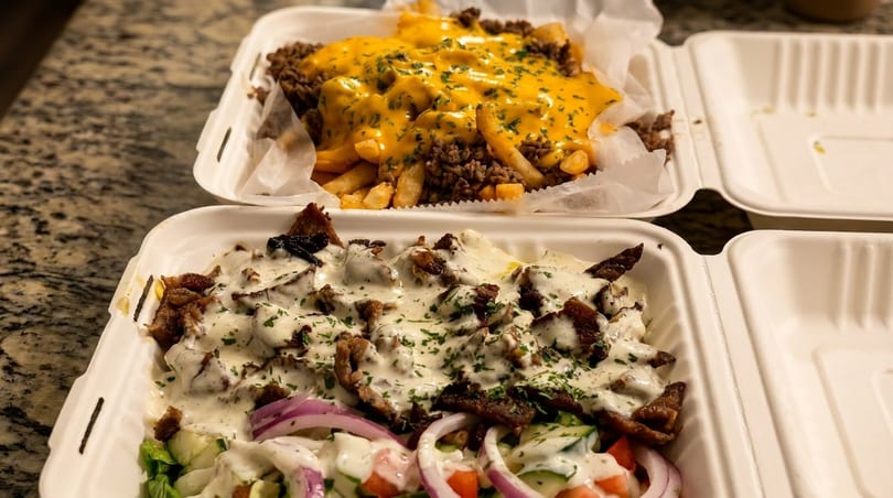 Two takeout containers with a cheesesteak fries and a gyro platter with white sauce.