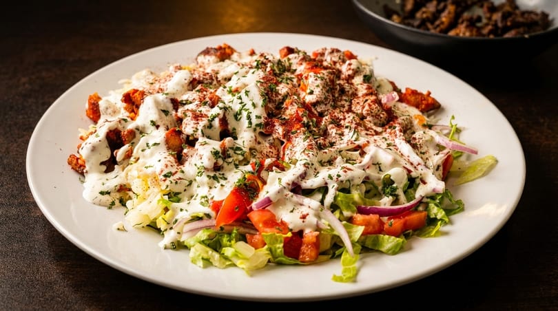A white plate of chicken and rice with salad, drizzled with white sauce and sprinkled with spices.