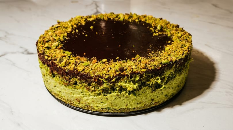 A round pistachio cheesecake with a dark chocolate ganache topping and crushed pistachios.